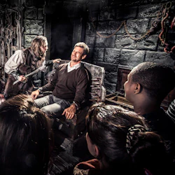 En man sitter i en tortyrstol och grimaserar, medan en annan man hotfullt håller ett verktyg nära honom, observerat av en grupp människor.