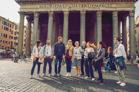 Group of tourists with audio guides standing in front of an ancient building with large columns and inscription.