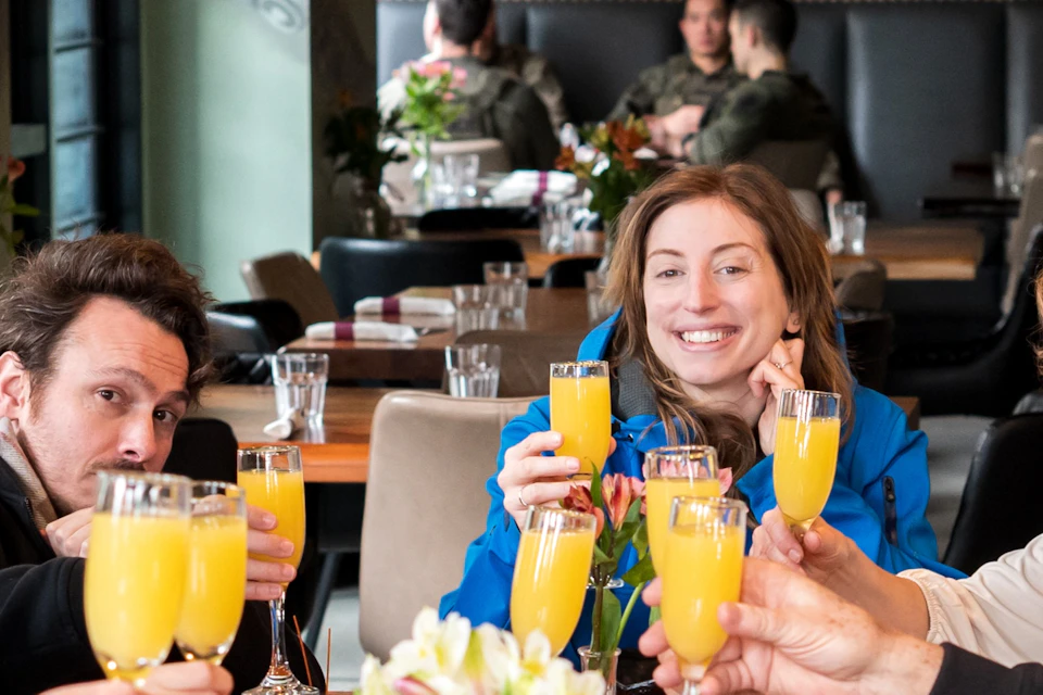 A group of smiling people sitting at a restaurant table, raising glasses filled with orange drinks in a toast.