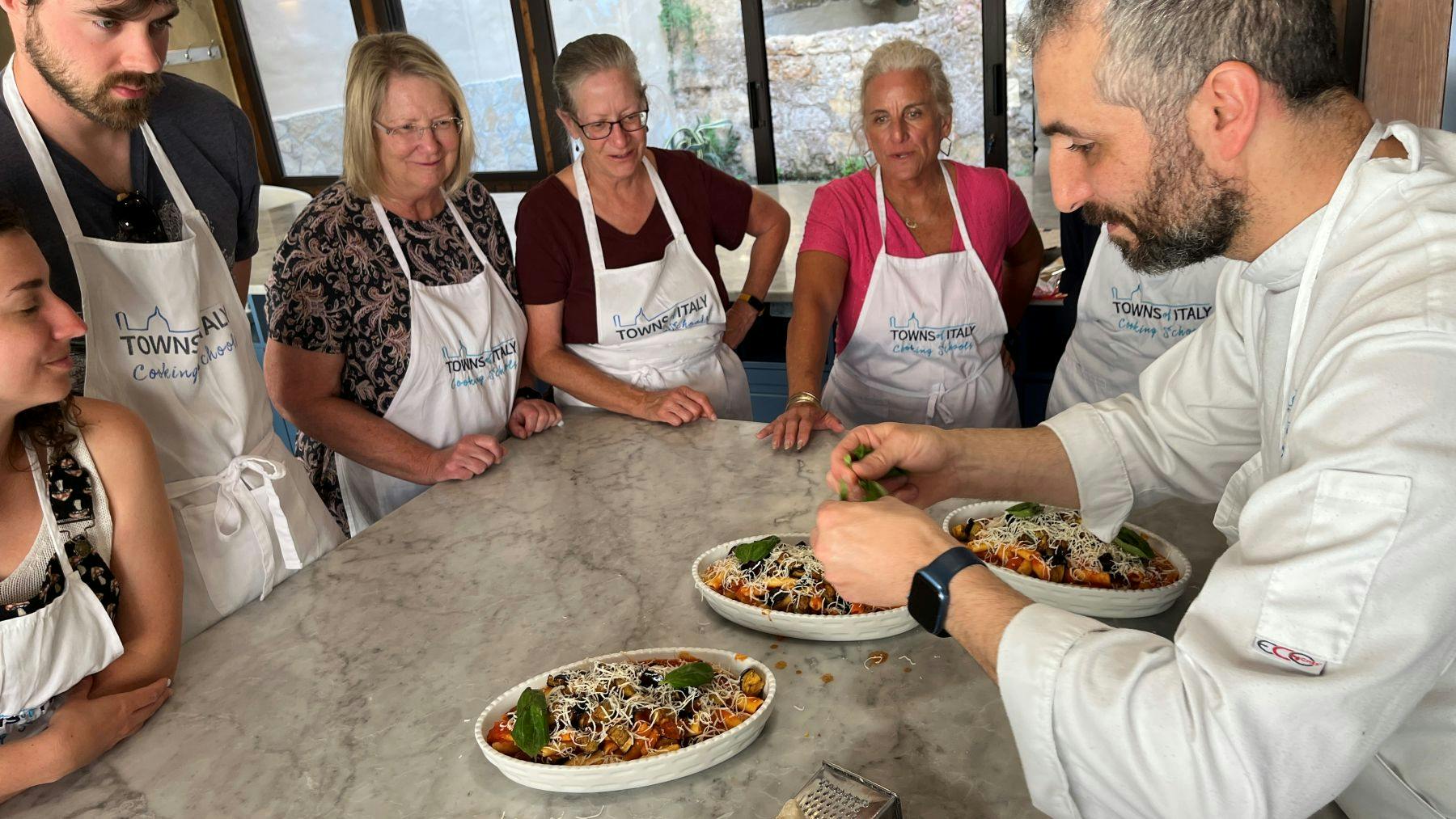 A group of people wearing aprons stand around a table with prepared dishes of food, observing as someone garnishes them.