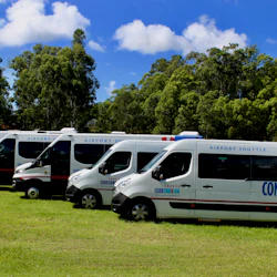 Four white airport shuttle vans parked on grass, with a backdrop of trees and a partly cloudy sky.