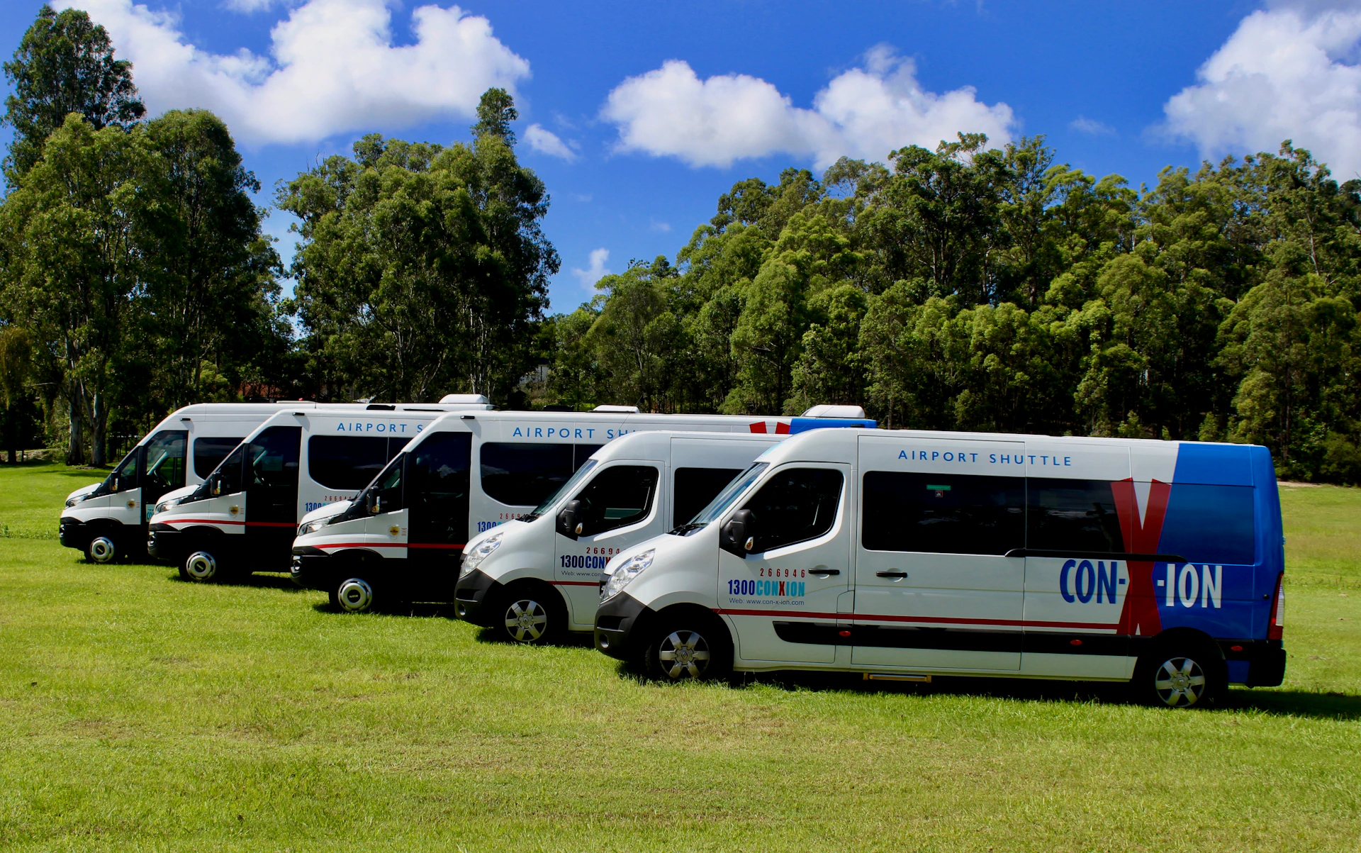 Four white airport shuttle vans parked on grass, with a backdrop of trees and a partly cloudy sky.