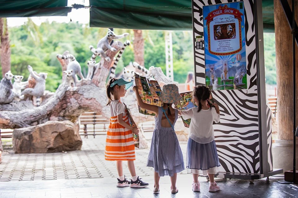 Three children stand in an outdoor area near a tiger show sign, holding and looking at a large map, with lemur statues behind.