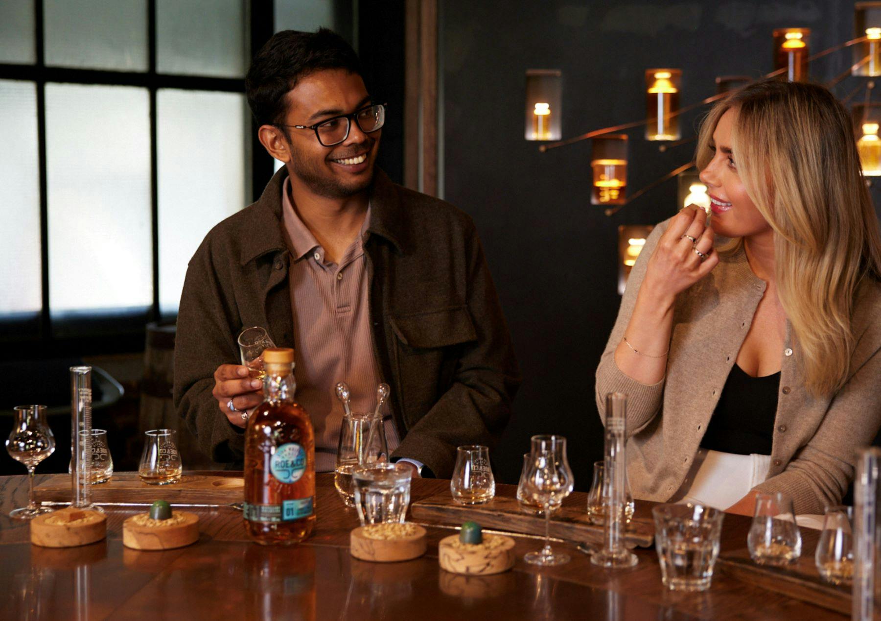 Muž se usmívá, zatímco drží sklenici whisky u stolu s různými skleněnými nádobami a lahví skotské whisky Highland Park Single Malt.