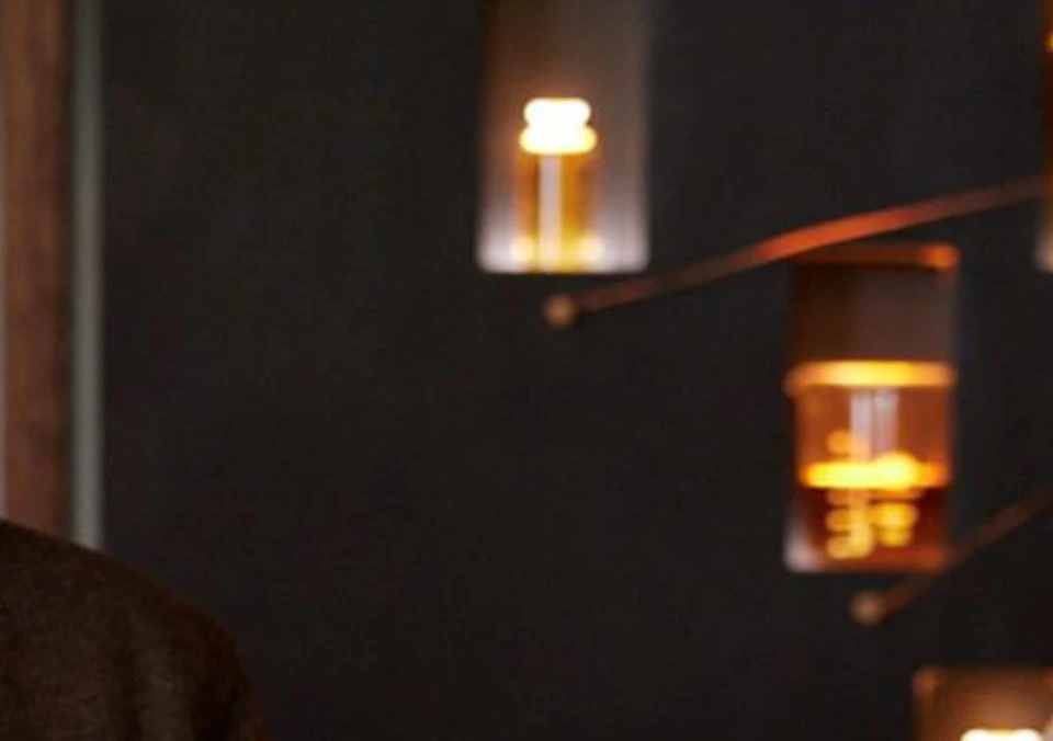 A man is smiling while holding a whiskey glass at a table with various glassware and a bottle of Highland Park Single Malt Scotch Whisky.