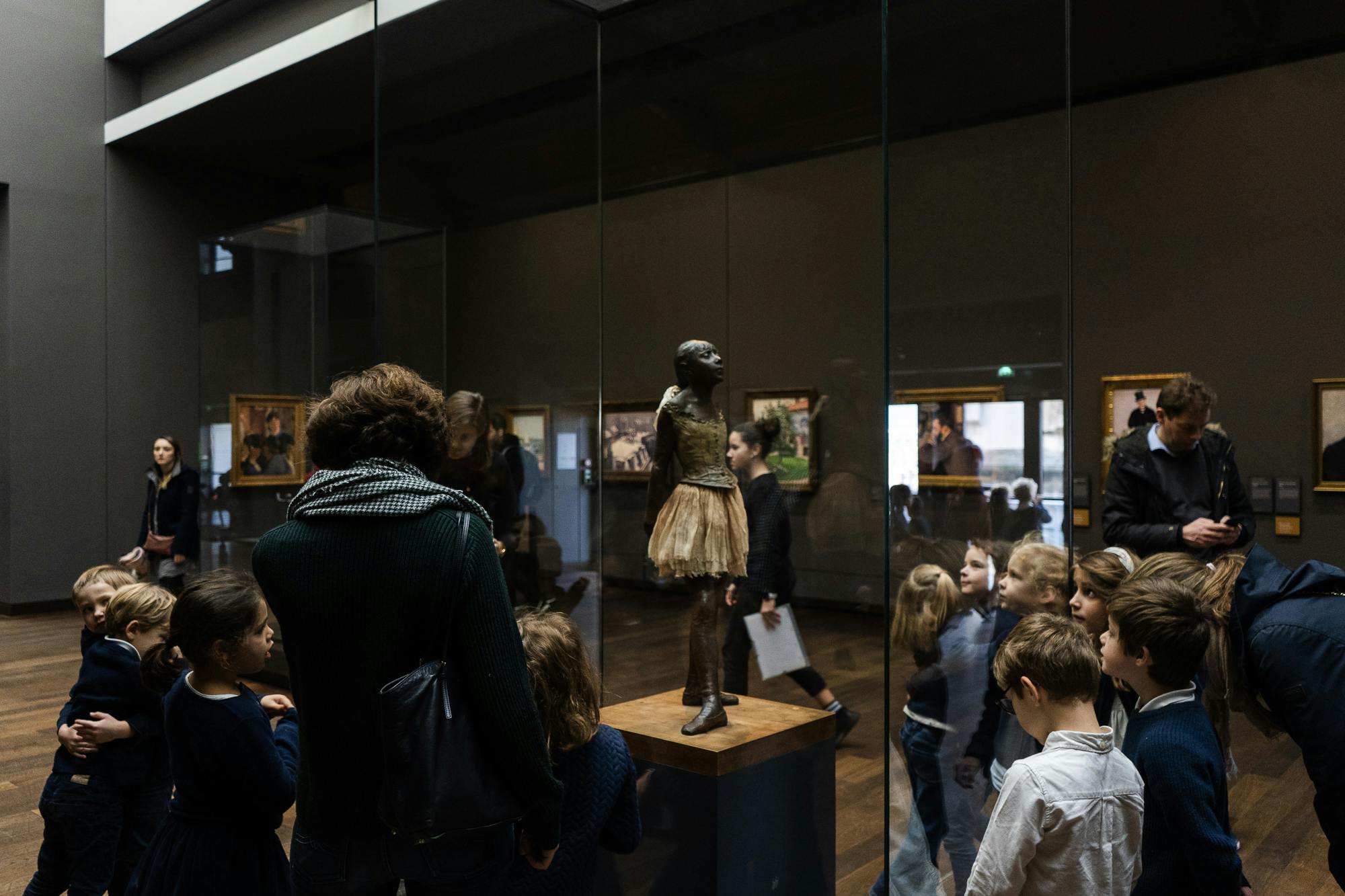 사람들은 박물관의 유리 케이스에서 청동 발레리나 동상을 관찰합니다. 미술품이 벽에 걸려 있습니다.