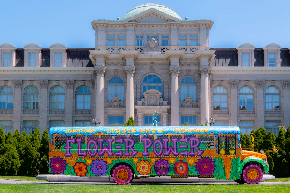 A brightly painted bus with "Flower Power" text and floral patterns parked in front of a historic building on a sunny day.