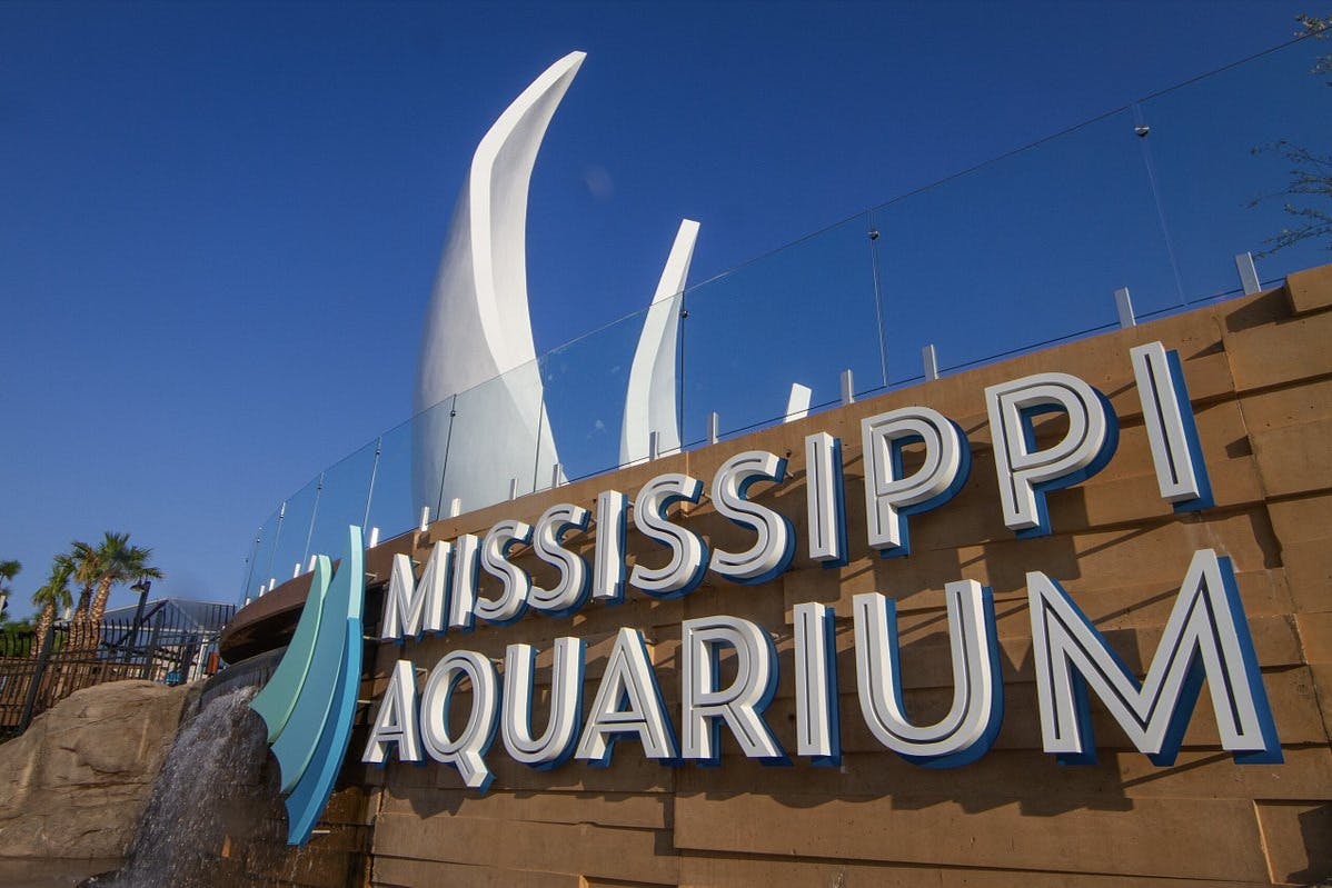 "Mississippi Aquarium" sign with modern architecture and water feature in front of a blue sky.