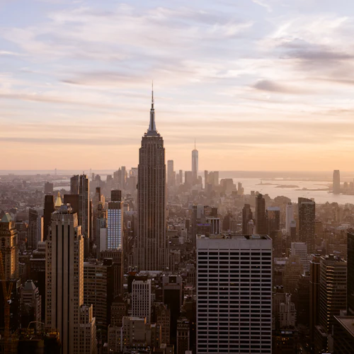 Blick auf eine Stadtsilhouette bei Sonnenuntergang, dominiert von hohen Wolkenkratzern, mit einem Fluss und entfernten Gebäuden im Hintergrund.