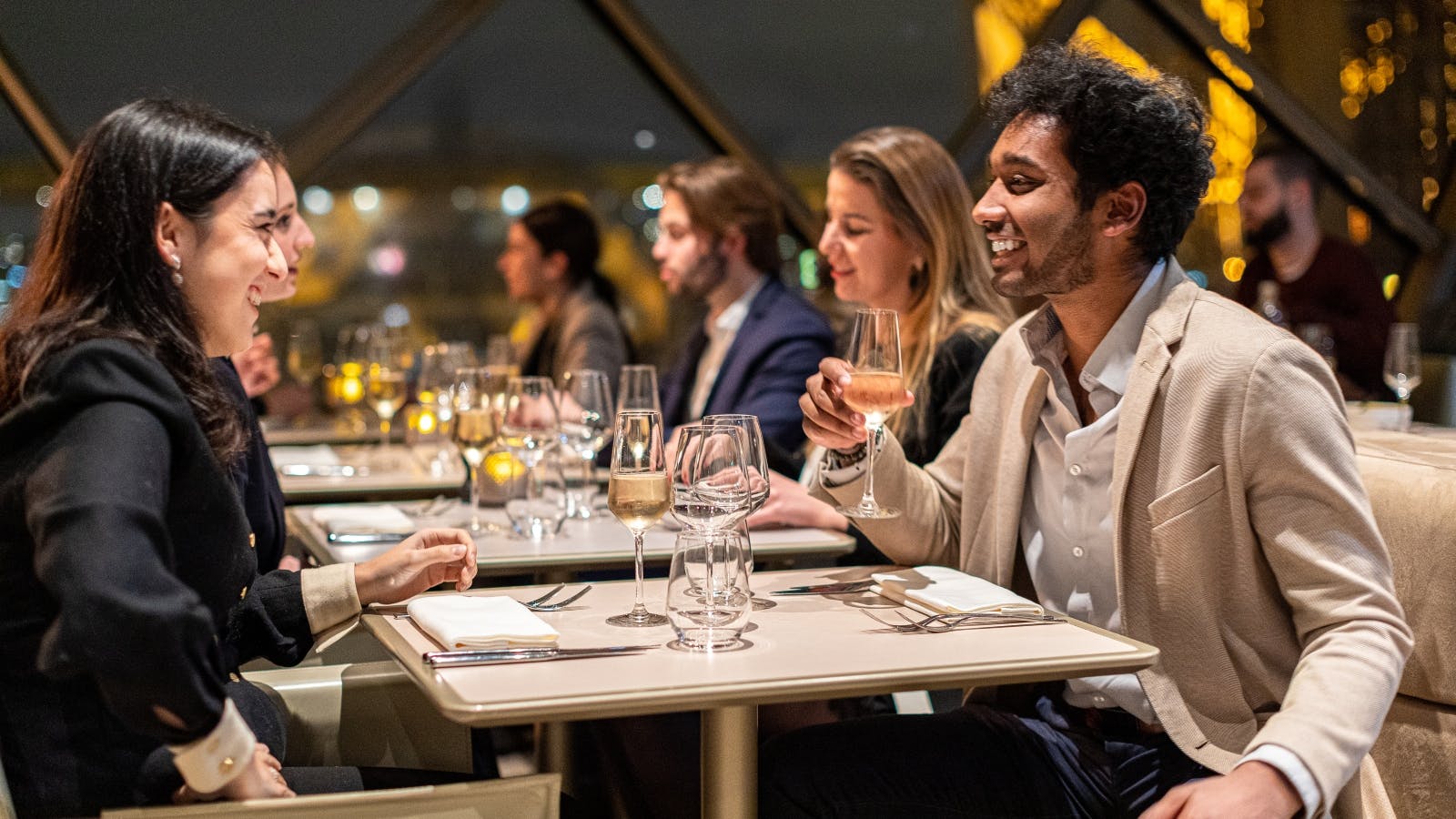 People dining at a restaurant, smiling while holding glasses of wine, with other diners visible in the background.