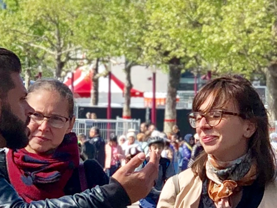 A group of six people standing outdoors on a sunny day with a busy street, green trees, and blue sky in the background.