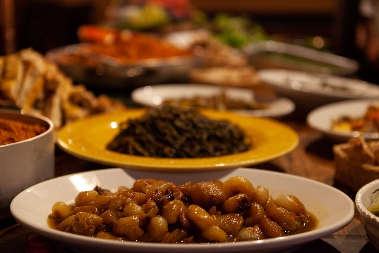 A variety of dishes on a table, including a bowl of cooked onions in the foreground and a plate of greens in the background.
