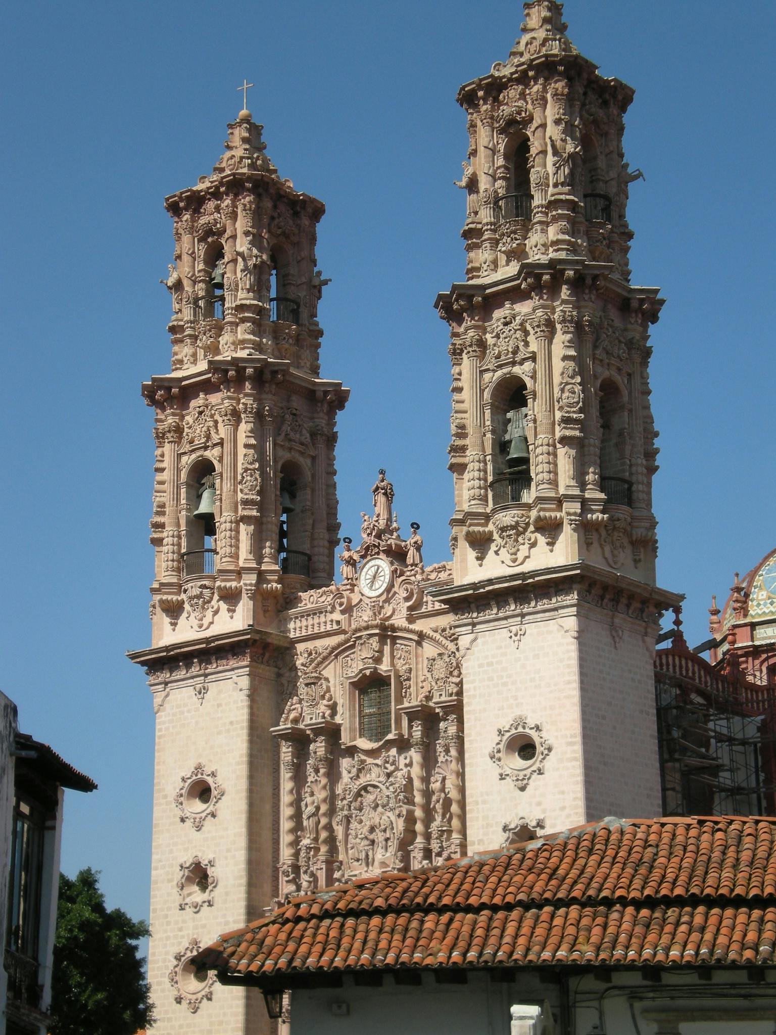 Verschnörkelte Zwillingsglockentürme eines historischen Gebäudes mit detaillierten Schnitzereien und einer Uhr, unter einem klaren blauen Himmel.