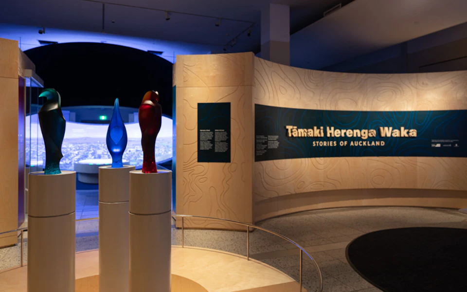 Exhibit space with a curved display reading 'Tāmaki Herenga: Stories of Auckland,' featuring glass sculptures in the foreground.