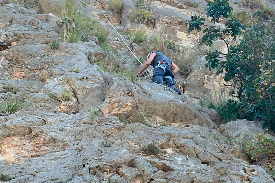 Person in climbing gear scaling a rocky cliff with vegetation, holding onto the rock and a rope for support.