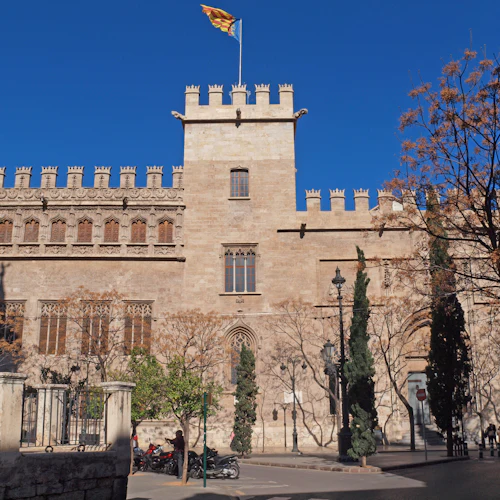 Historisk stenbyggnad med en krenelerad takterrass, gotiska detaljer och flagga på toppen. Träd och människor framför. Klarblå himmel.