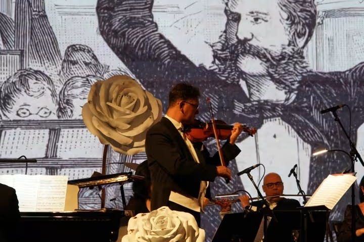 En violinist optræder sammen med et orkester med en stor vintage tegning af en mand med et overskæg i baggrunden.