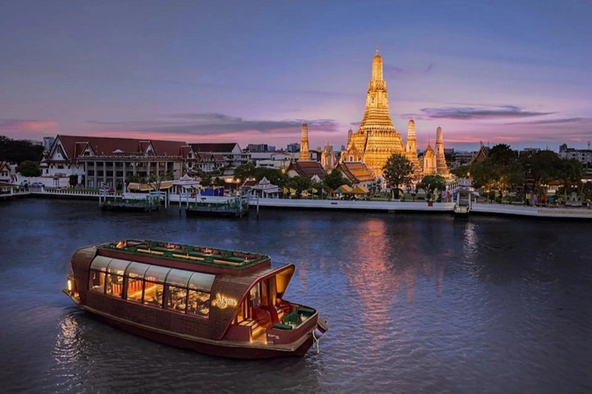 A boat on a river at sunset with a lit-up temple in the background, surrounded by other buildings and trees.