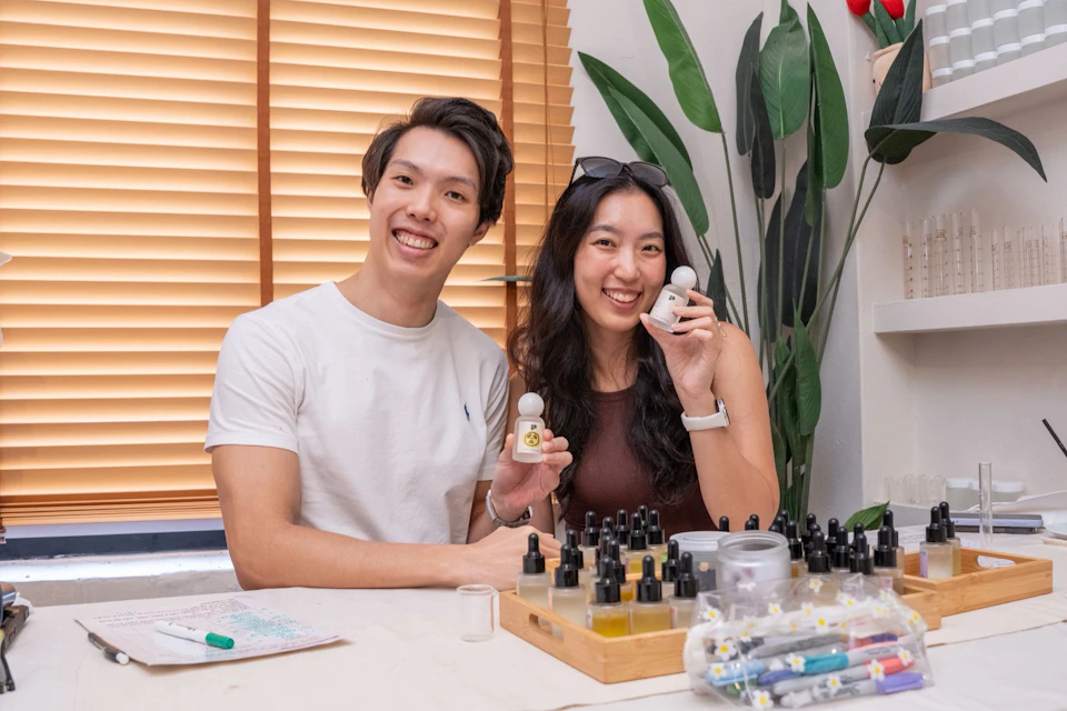 Two people sitting at a table with small bottles and tools, smiling and holding items, with a plant and window blinds behind them.