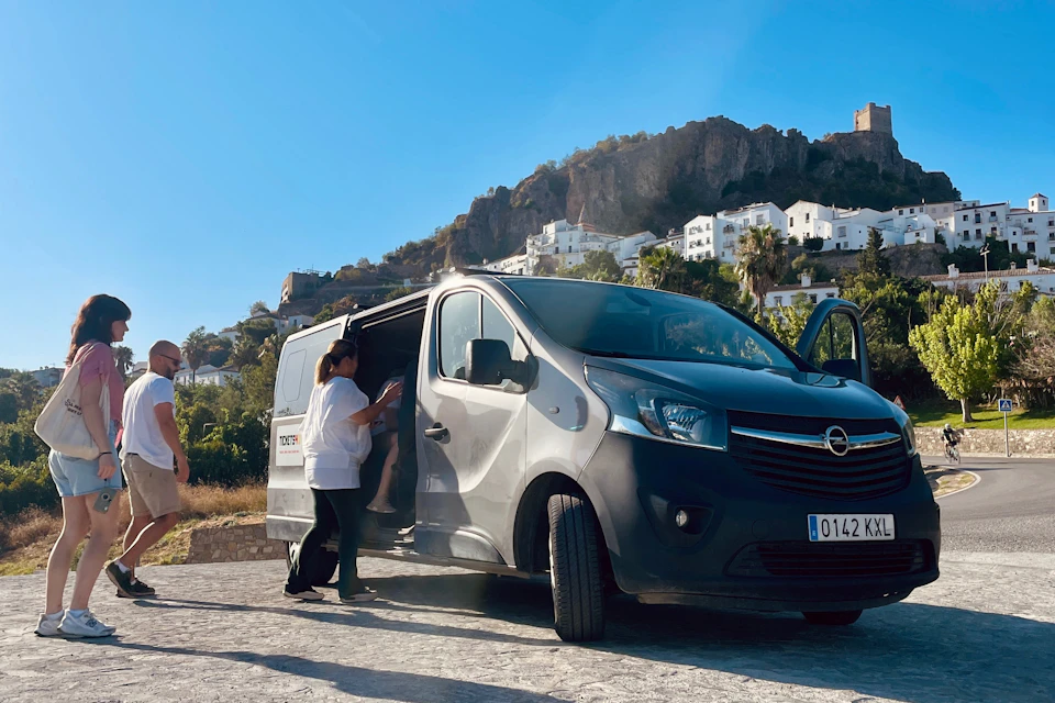 A person helps another into a black van in a sunny outdoor setting with white buildings and a rocky hill in the background.