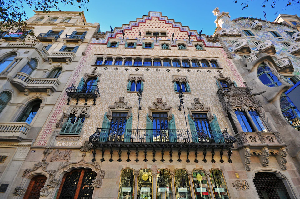 Ornate building facade with intricate patterns, wrought-iron balconies, and large windows, flanked by adjacent buildings.