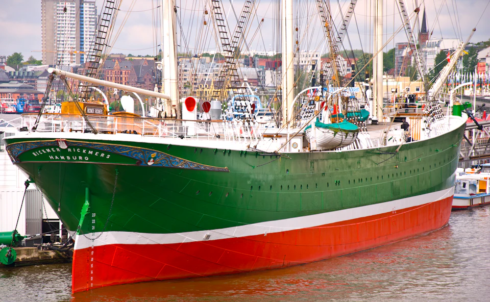 A large green and red ship docked at a harbor with buildings and trees in the background under a cloudy sky.