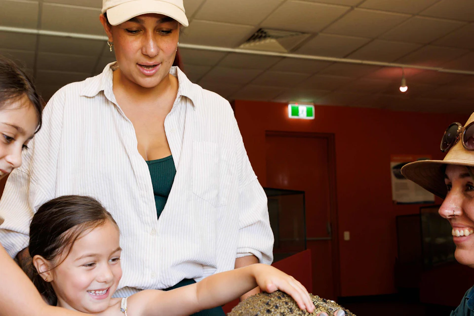 Two children pet a small crocodile held by a smiling woman, accompanied by another adult, indoors.