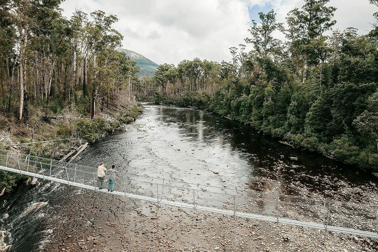 Two people stand on a suspension bridge over a wide river surrounded by lush green forest under a cloudy sky.