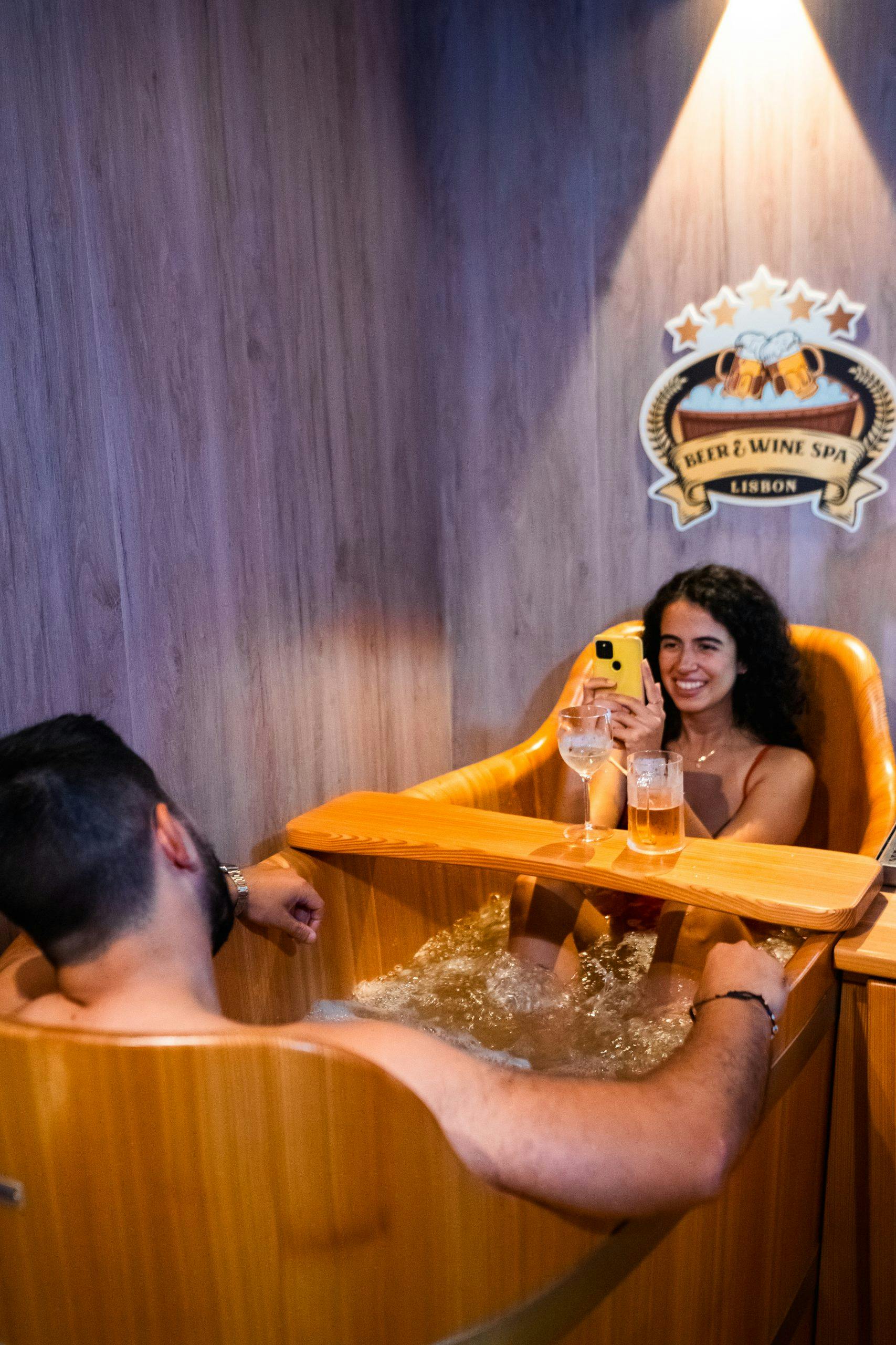 Δύο άτομα χαλαρώνουν σε ένα ξύλινο υδρομασάζ σε ένα σπα, με ποτά και ένα τηλέφωνο. Η ταμπέλα στον τοίχο γράφει "Beer & Wine Spa Lisbon."