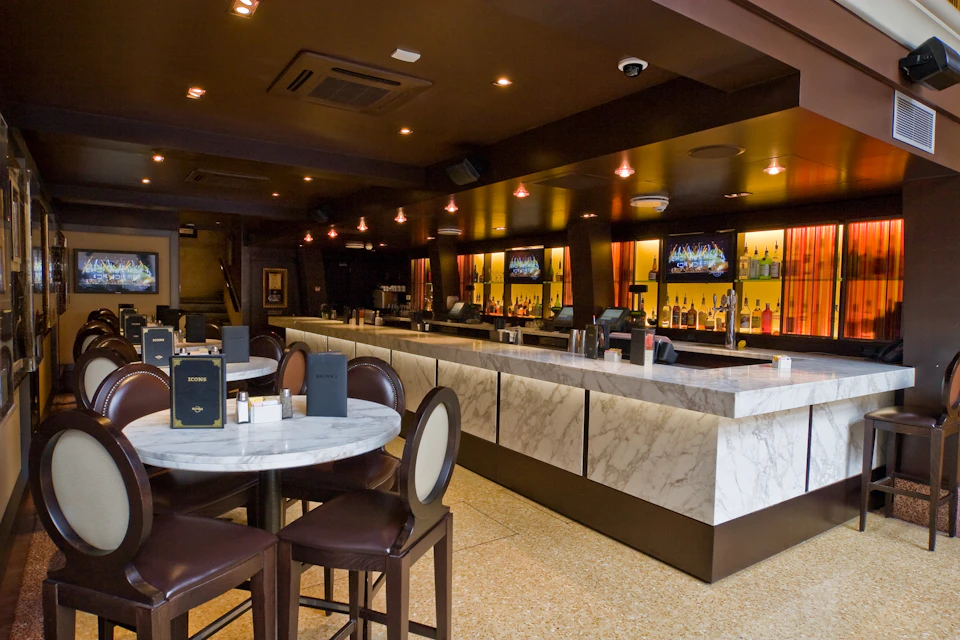 Modern bar with marble countertops, illuminated shelves with bottles, cozy round tables with chairs, and two wall-mounted TVs.
