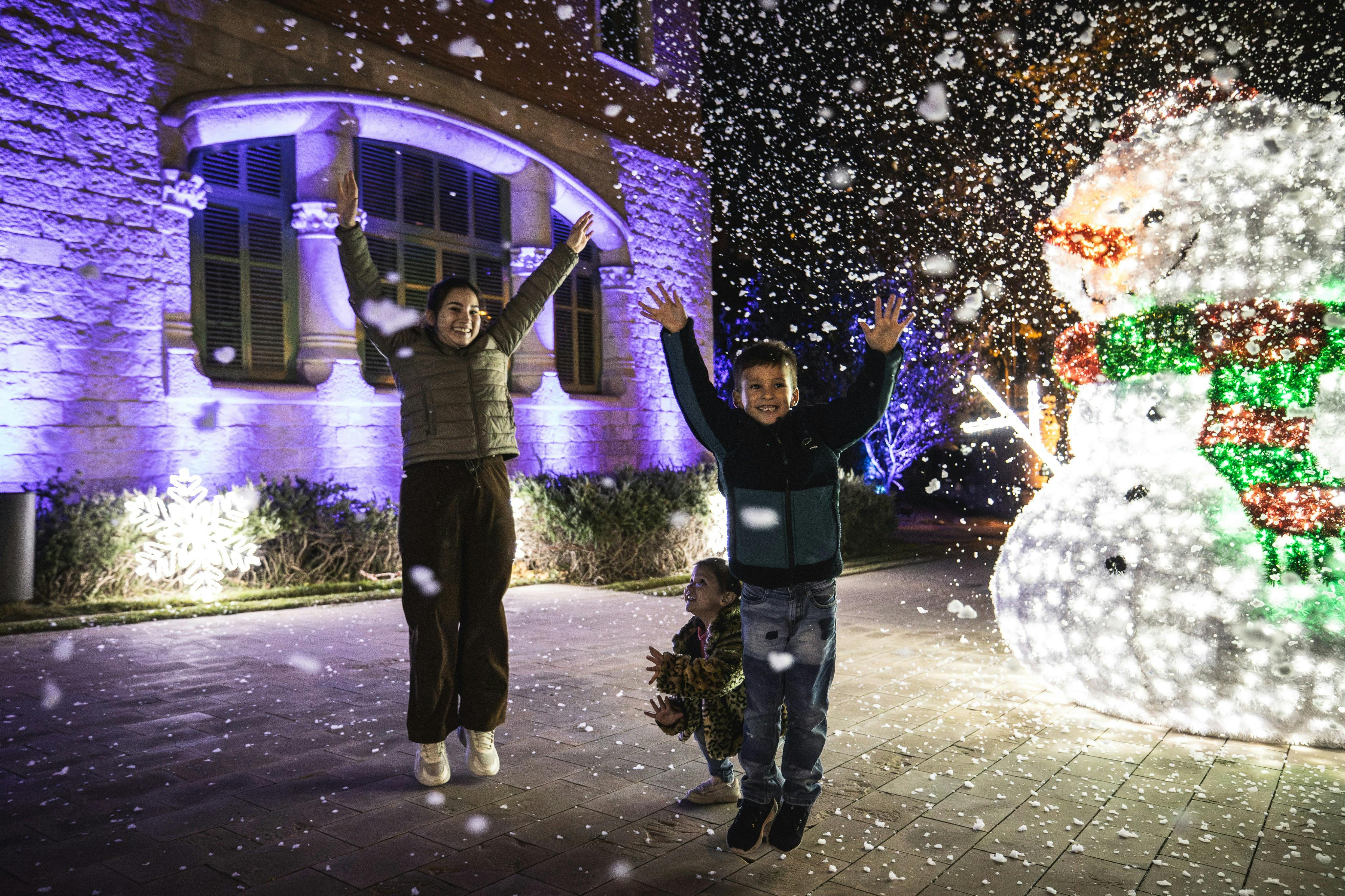 Three children are joyfully playing in falling snow in front of a lit building at night.