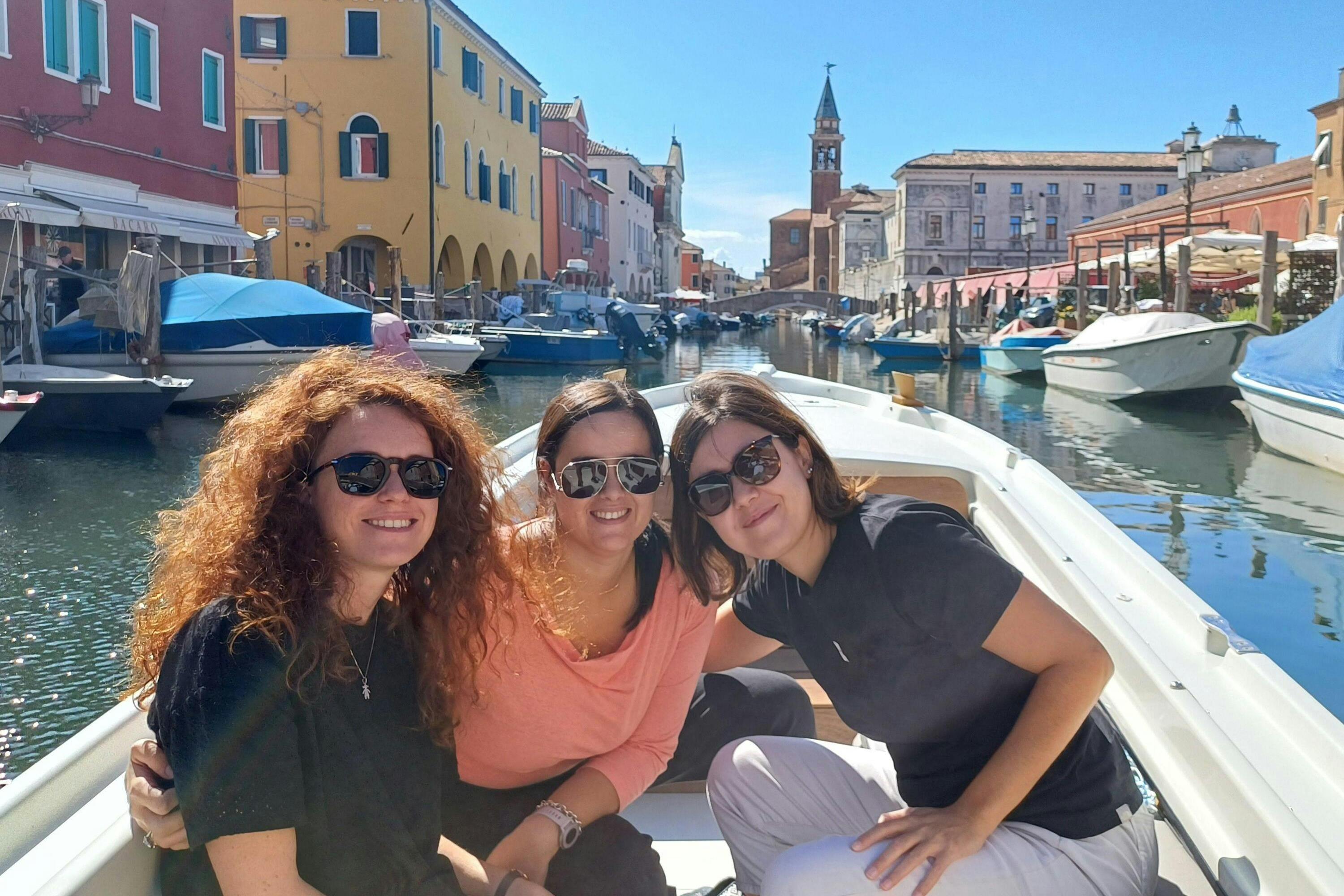 Three women wearing sunglasses sit on a boat in a canal with colorful buildings and a bell tower in the background.