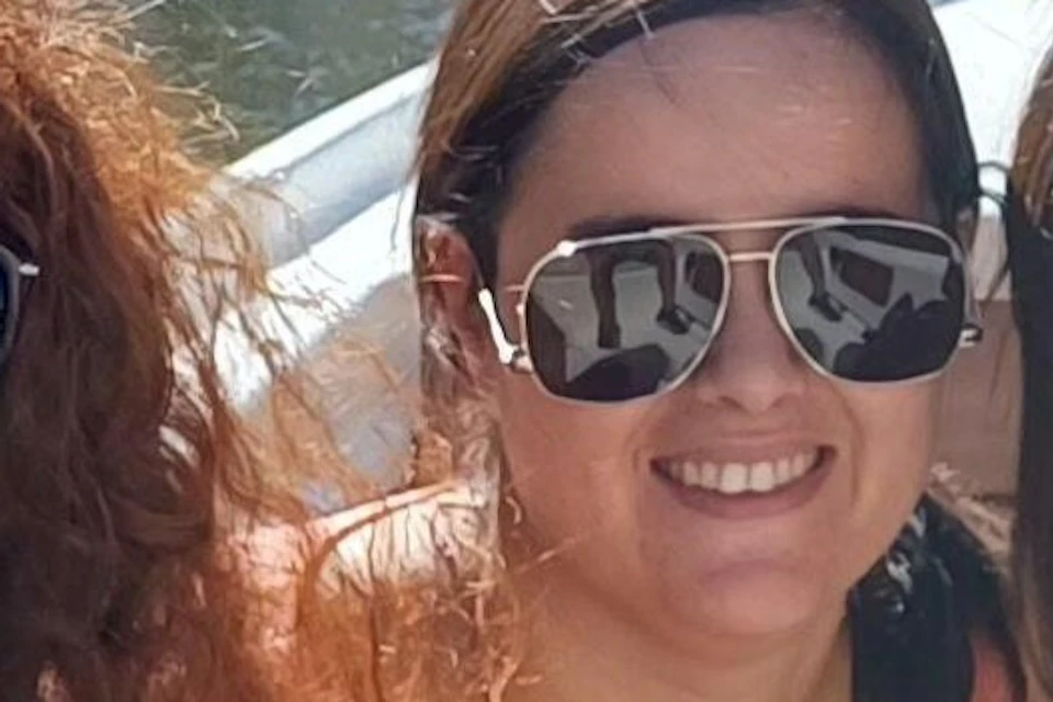 Three women wearing sunglasses sit on a boat in a canal with colorful buildings and a bell tower in the background.