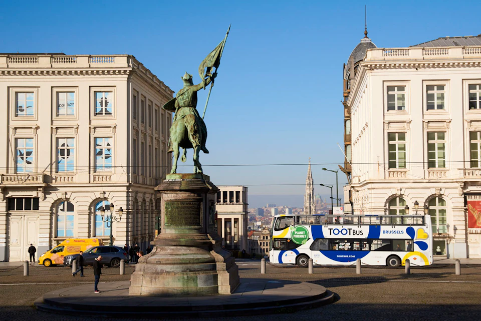 Equestrian statue in a city square flanked by two ornate buildings, with a tour bus and pedestrians in the background.