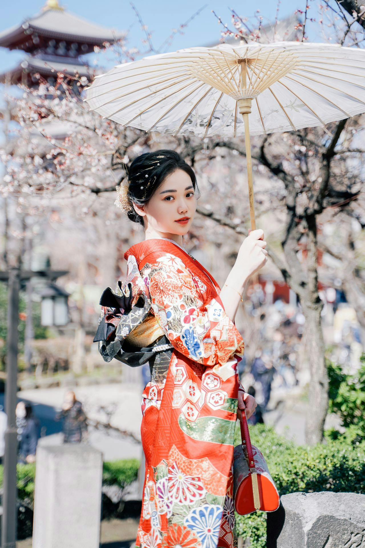 A person in a traditional colorful kimono holds a parasol in a garden with blooming trees and a pagoda in the background.