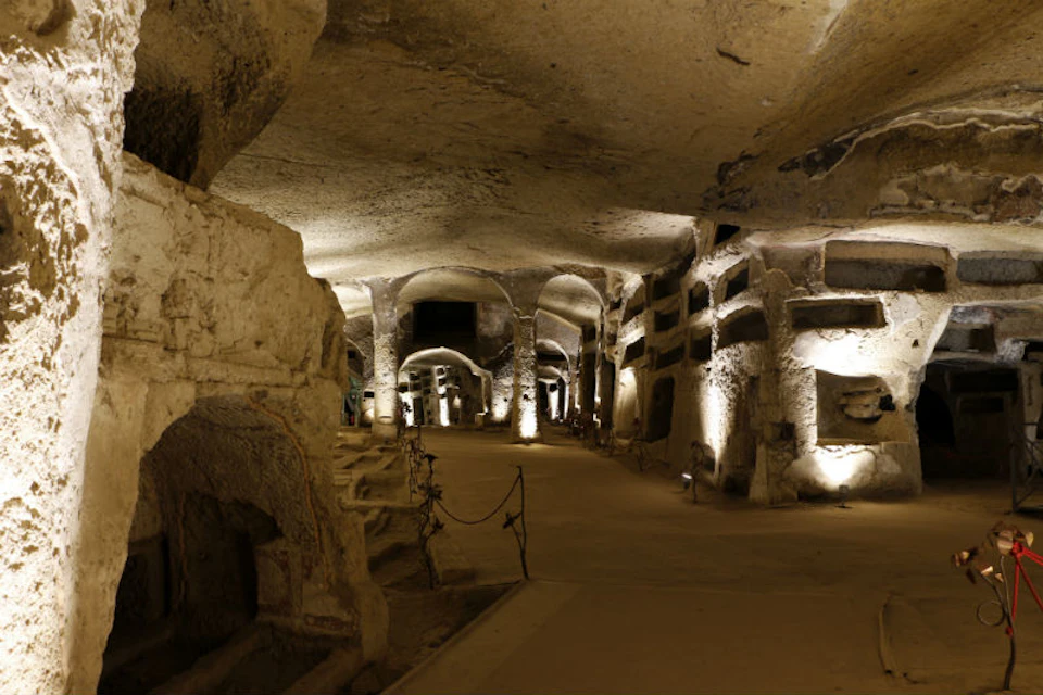 An illuminated underground catacomb with stone arches, niches, and pathway.