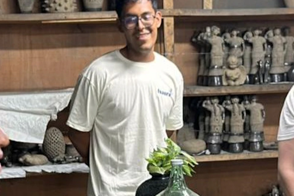 Three individuals stand around a wooden table in a workshop with pottery displayed on shelves in the background.