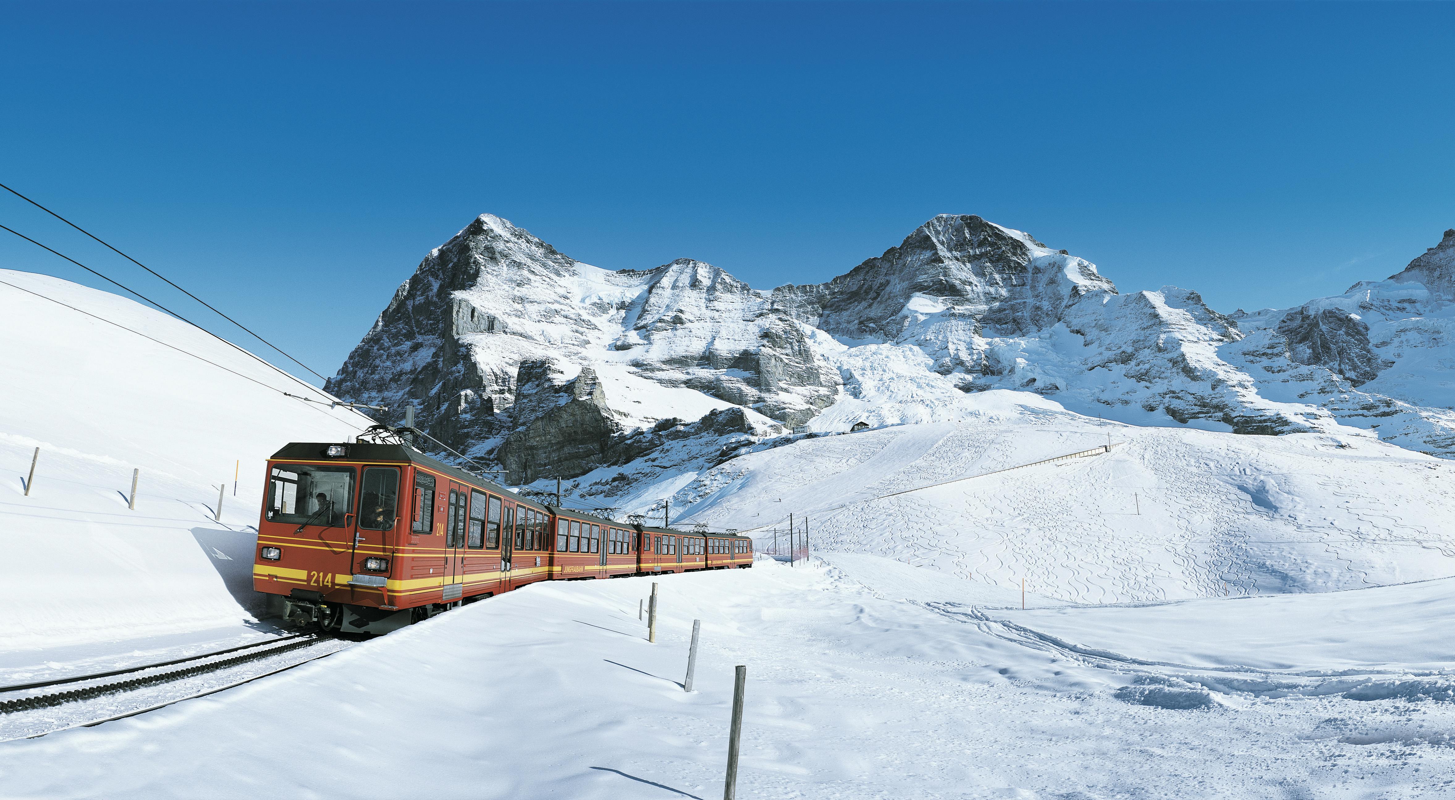 Červený vlak projíždí zasněženou hornatou krajinou s jasně modrou oblohou a zasněženými vrcholky.