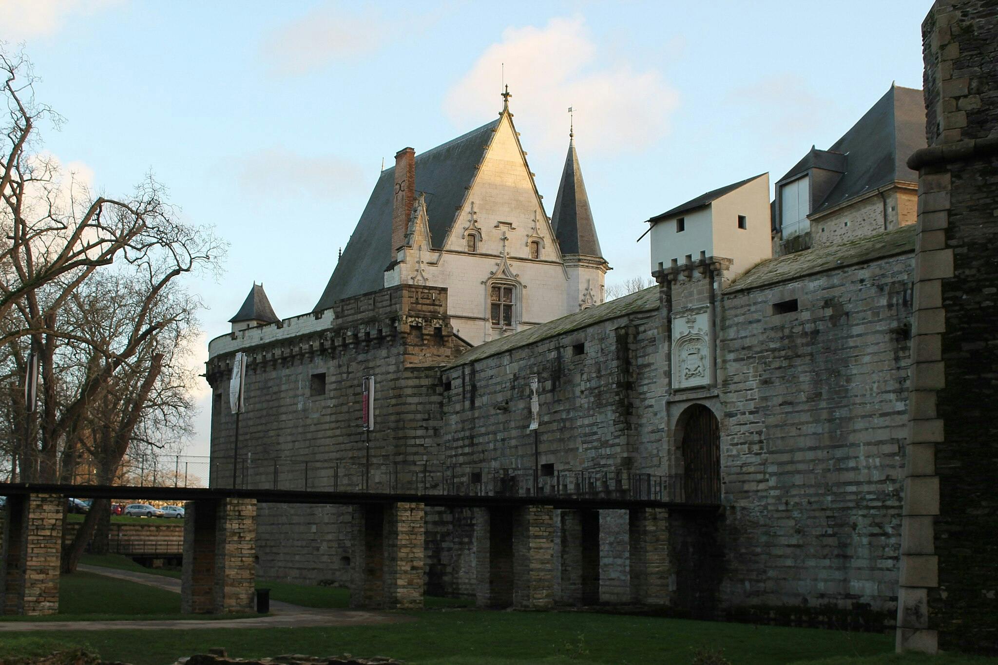 Château en pierre avec un grand mur, une porte voûtée et des tourelles pointues, entouré de pelouse et d'arbres sous un ciel bleu.
