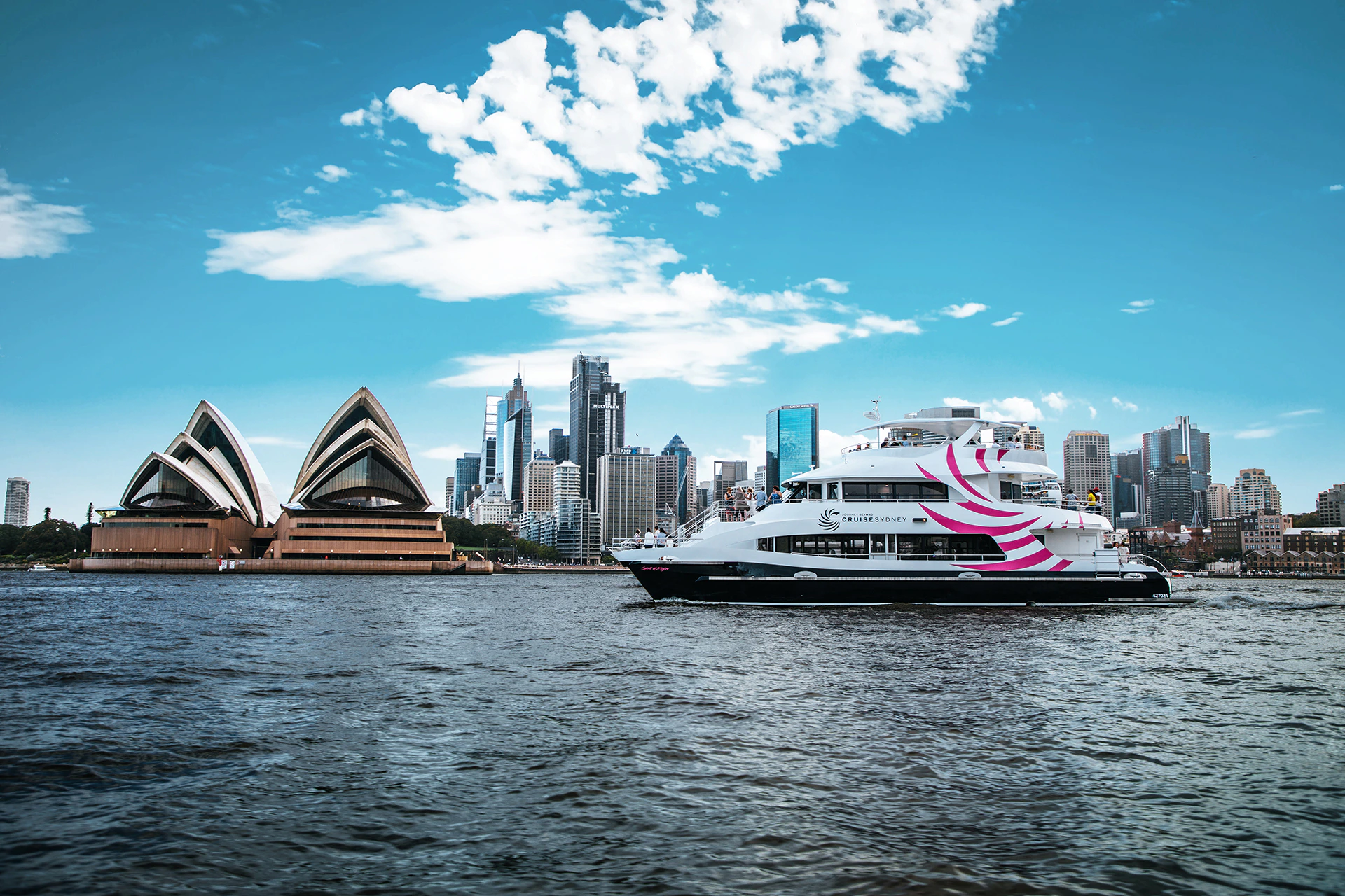 A boat navigates a harbor with the Sydney Opera House and a city skyline in the background under a partly cloudy sky.