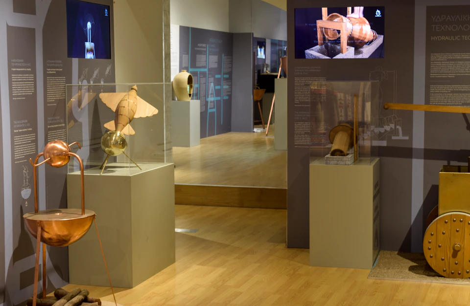 Museum exhibit featuring wooden models displayed in glass cases on pedestals, with informational panels and a floor mirror in the background.