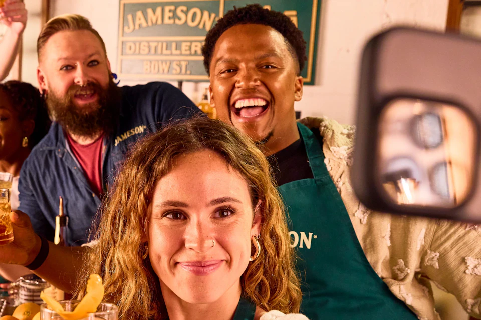 A group of people behind a bar smiling and holding drinks while one takes a selfie with a smartphone.