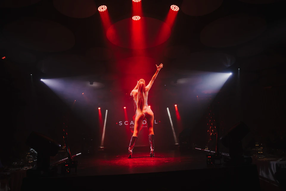 Performer on stage under red and white lights, with arms raised, in a dramatic pose during a show.