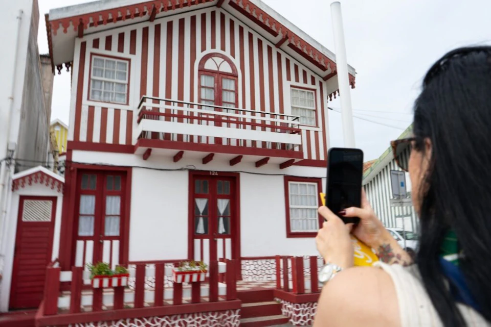 A person takes a photo of a white and red striped house with a small balcony and red doors.