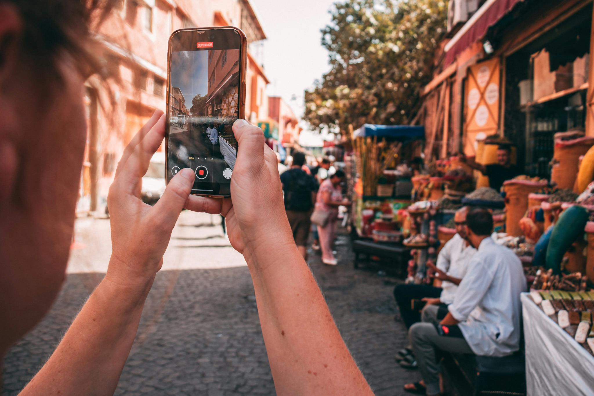 Ένα άτομο που κρατάει ένα smartphone, φωτογραφίζει μια πολυσύχναστη υπαίθρια αγορά με ανθρώπους, πολύχρωμους πάγκους και ηλιόλουστα κτίρια.