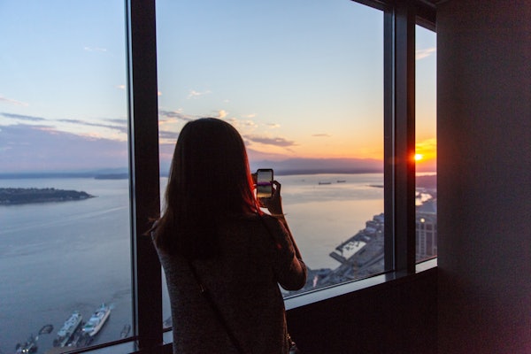 高層ビルの大きな窓から夕日の写真を撮っている女性。