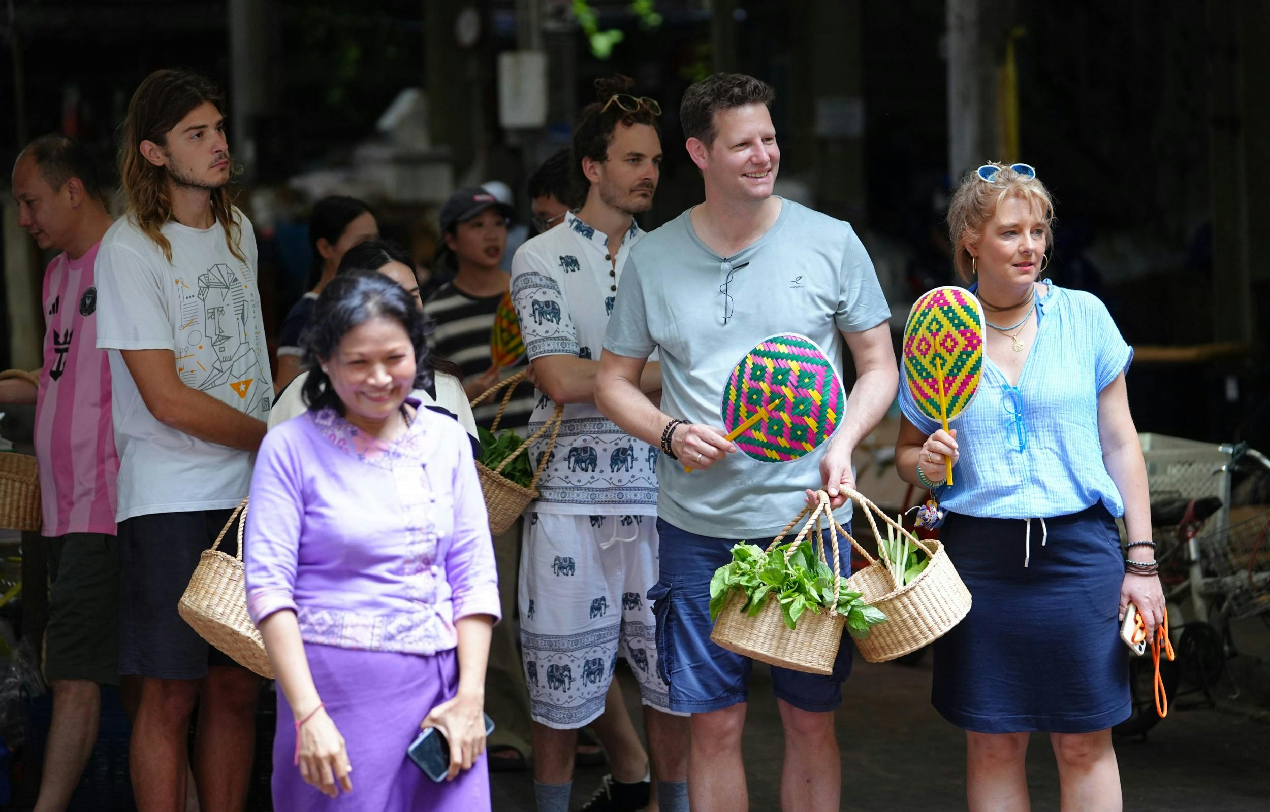 A group of people standing outdoors holding woven baskets with greens, one person holding a colorful hand fan.