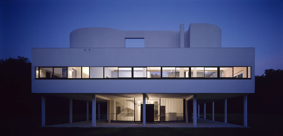 Modern, minimalist white building with large windows, elevated on pillars, and illuminated at dusk against a deep blue sky.