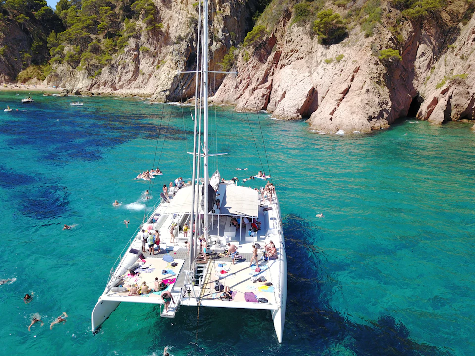 A catamaran anchored in clear blue water near rocky cliffs, with people swimming and sunbathing on the boat.