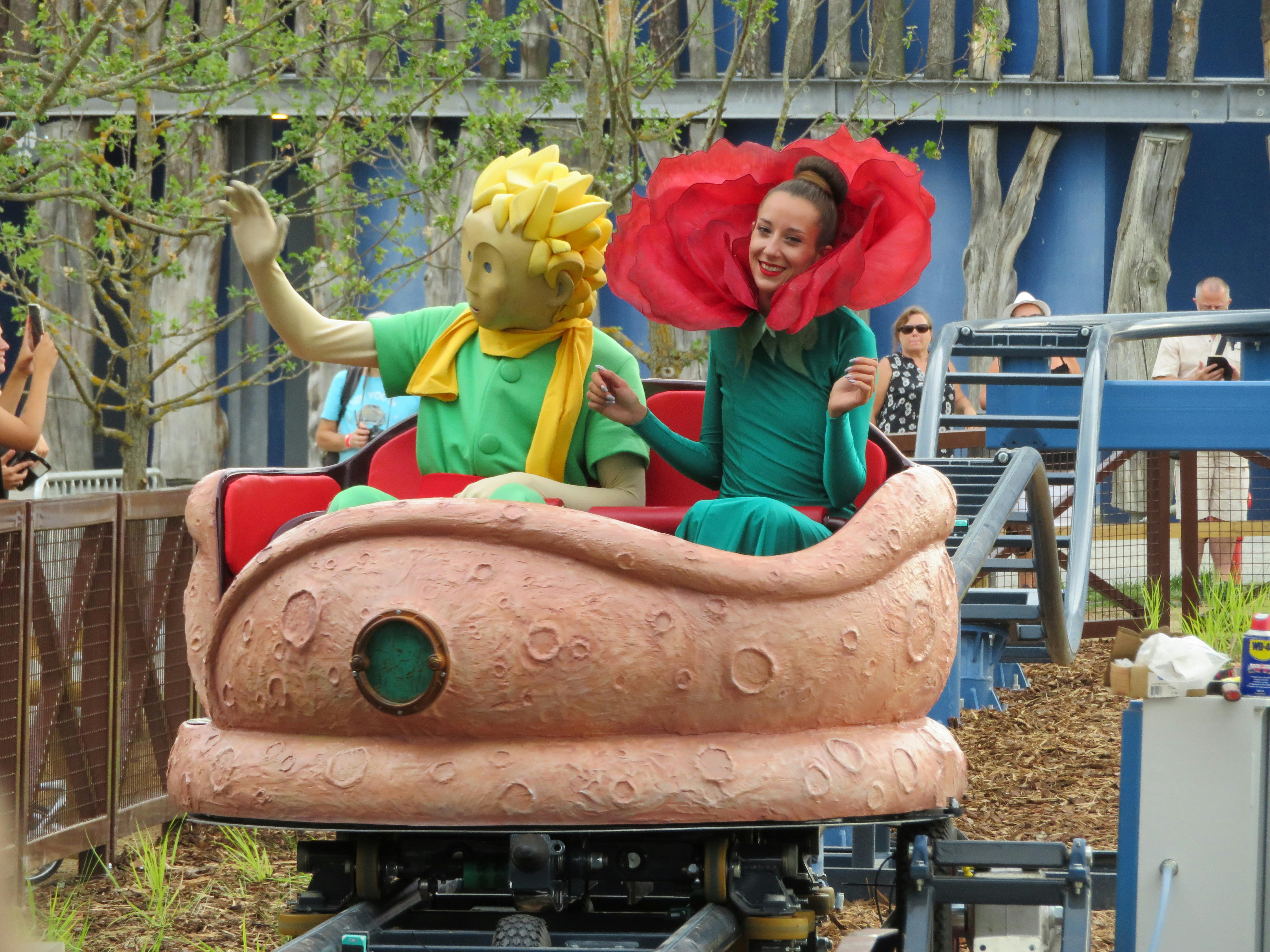 꽃 의상을 입은 사람과 초록색과 노란색 의상을 입은 인형이 놀이공원에서 기발한 놀이기구에 앉아 있습니다.